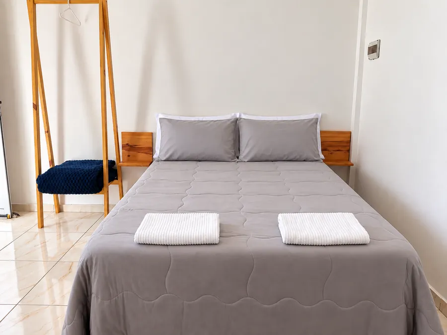 Cama matrimonial gris con almohadas grises, toallas blancas y perchero de madera